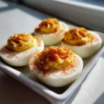 Four perfectly piped classic deviled eggs sprinkled generously with bright red paprika on a white dish.