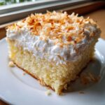 A close-up slice of moist coconut poke cake topped with whipped cream and toasted coconut flakes.