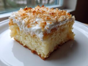 A close-up of a moist slice of coconut poke cake topped with whipped cream and toasted coconut flakes.
