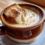Close-up of a piece of toasted bread dipping into a rich, creamy cheese fondue served in a brown crock.