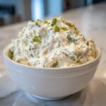 A white bowl filled with creamy clam dip, garnished generously with chopped green onions or chives.