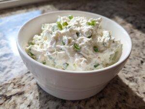 A white bowl filled with creamy clam dip, garnished with chopped green onions, sitting on a granite countertop.