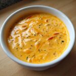 A close-up of rich, orange cottage cheese queso dip topped with herbs and red pepper pieces in a white bowl.
