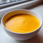 Close-up of a bowl filled with vibrant orange, creamy cheese soup, sitting on a white surface.