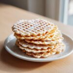 A tall stack of freshly made, golden brown pizzelle cookies dusted generously with powdered sugar on a white plate.