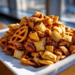 A close-up, appetizing photo of a mound of seasoned chex party mix, including square cereal pieces and pretzels, piled high on a white dish.