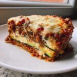 A thick, firm slice of zucchini lasagna featuring layers of meat sauce, ricotta, and zucchini noodles, topped with melted cheese.