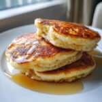 A close-up of a stack of three fluffy banana pancakes generously drizzled with golden syrup on a white plate.