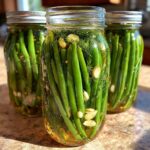 Three glass jars filled with bright green dilly beans, fresh dill, and garlic cloves, ready for pickling.
