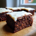 A close-up, appetizing shot of a square slice of dark, moist gingerbread brownies topped with thick white frosting.