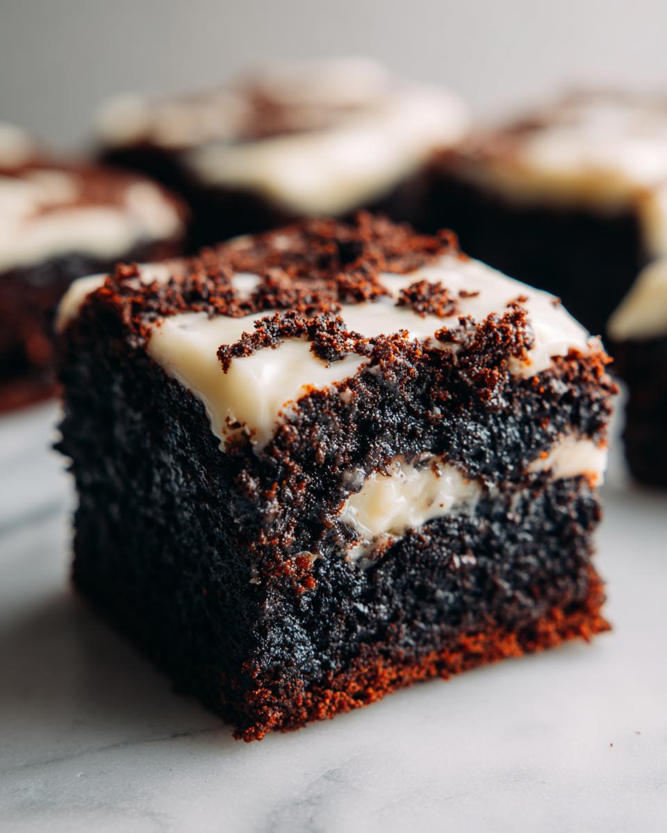 A close-up shot of a rich, dark square of gingerbread brownies topped with white frosting and chocolate crumbs.