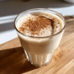 A glass of creamy, frothy coquito topped generously with ground cinnamon, sitting on a wooden surface.