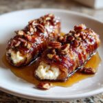 Two bacon-wrapped stuffed dates, glazed with caramel and topped with chopped pecans, served on a white plate.