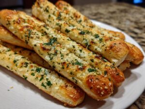 Close-up of several golden brown, freshly baked breadsticks seasoned with garlic and parsley on a white plate.