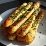 A stack of freshly baked, golden brown breadsticks seasoned with garlic butter and sprinkled generously with chopped parsley.