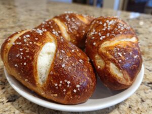 Three freshly baked pretzel dogs buns, golden brown and topped with coarse salt, resting on a white plate.
