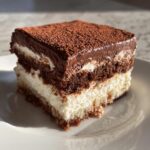 A square slice of rich, layered Italian love cake featuring white cream, dark chocolate cake, and a cocoa powder topping.