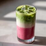 Close-up of a tall glass containing a layered strawberry matcha drink with ice cubes, showing pink, white, and green layers.