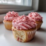 Close-up of moist strawberry cupcakes with fluffy pink strawberry frosting; one has a bite taken out.