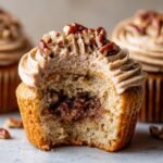 Close-up of a pecan pie cupcakes with a bite taken out, showing the gooey filling, spiced frosting, and pecan topping.