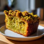 A close-up, sunlit slice of pistachio coffee cake showing a vibrant green interior and a thick, crunchy pistachio crumb topping.