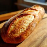A freshly baked loaf of golden brown french bread resting on a wooden cutting board, showing a crisp, scored crust.