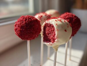 Close-up of several red velvet cake pops, one of which is partially dipped in white chocolate and shows the vibrant red interior.