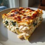 A generous slice of baked chicken alfredo lasagna showing layers of shredded chicken, spinach, and a bubbly, browned cheese topping.