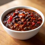 A close-up of a white bowl filled with rich, dark red black bean chili, featuring visible black beans and chunks of red pepper.