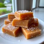 A stack of soft sea salt caramels sprinkled with coarse sea salt, sitting on a white plate.