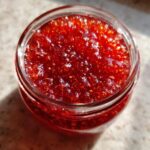 Top-down view of vibrant, chunky strawberry freezer jam filling a clear glass jar, glistening in the sunlight.