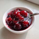 A spoonful of glossy, deep red cherry pie filling being lifted from a small white bowl.