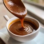 Close-up of thick homemade brown gravy being poured from a spoon into a white bowl.