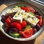A vibrant bowl of authentic greek salad featuring large blocks of feta cheese drizzled with oil and herbs, mixed with tomatoes, cucumbers, and Kalamata olives.