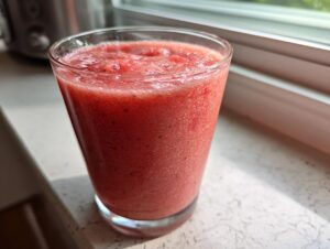 A tall glass filled with a vibrant, icy red watermelon smoothie sitting on a sunlit windowsill.