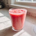 A tall glass filled with a vibrant pink, icy watermelon smoothie sitting on a bright white marble countertop.