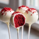Close-up of four red velvet cake pops dipped in white chocolate, one is cut open revealing the red interior.