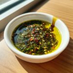 Close-up of a white bowl containing bright green herbs, chili flakes, and seeds submerged in golden olive oil bread dip.