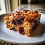 Close-up of a square slice of blueberry baked oatmeal on a white plate, showing a moist texture and caramelized top.