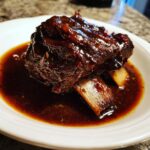 A single, perfectly cooked portion of braised short ribs covered in a dark, glossy sauce, served in a white bowl.