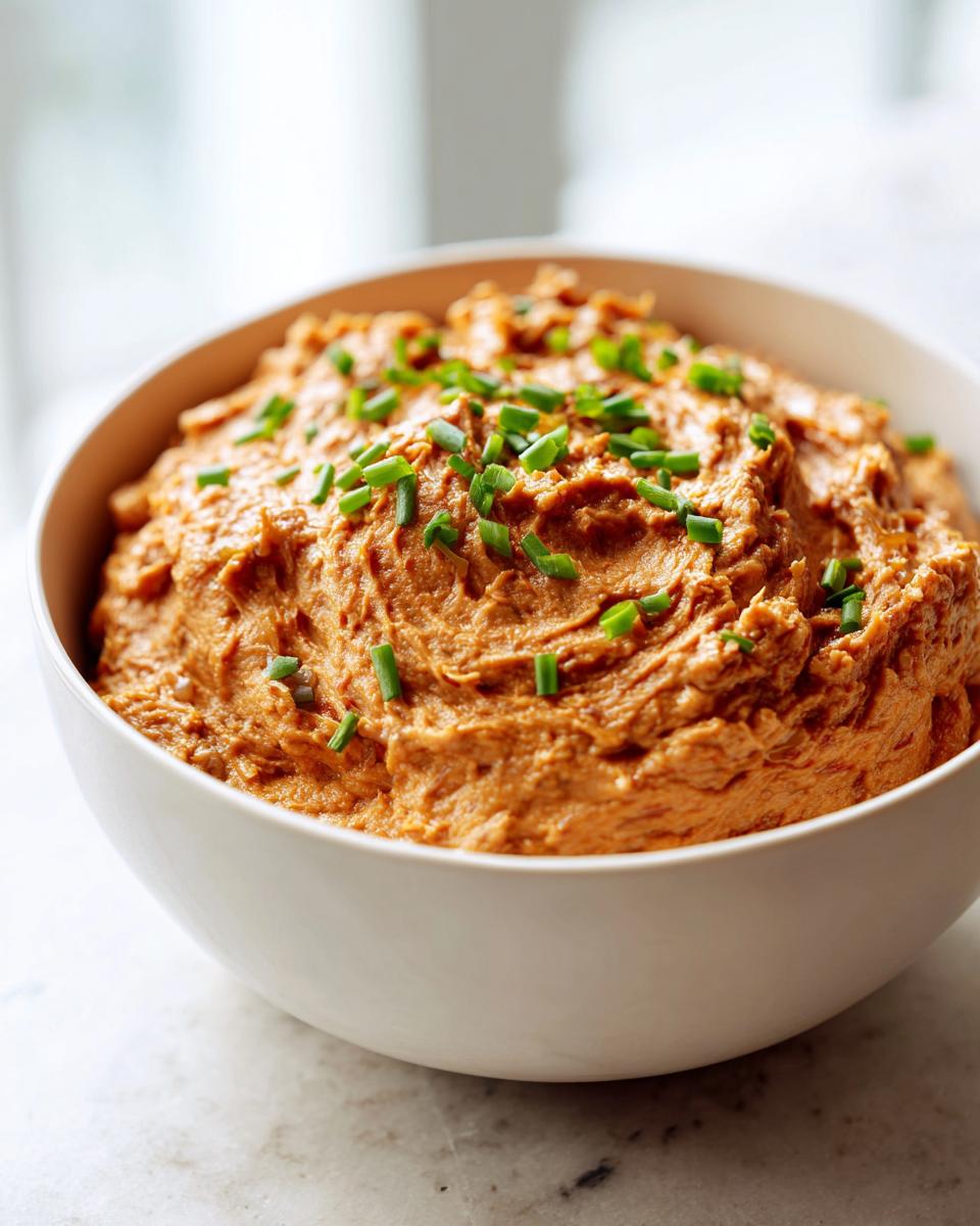 A creamy, rich orange-brown caramelized onion dip garnished with fresh chopped chives in a white bowl.