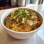 A close-up of creamy caramelized onion dip topped with fresh chives in a white serving bowl.