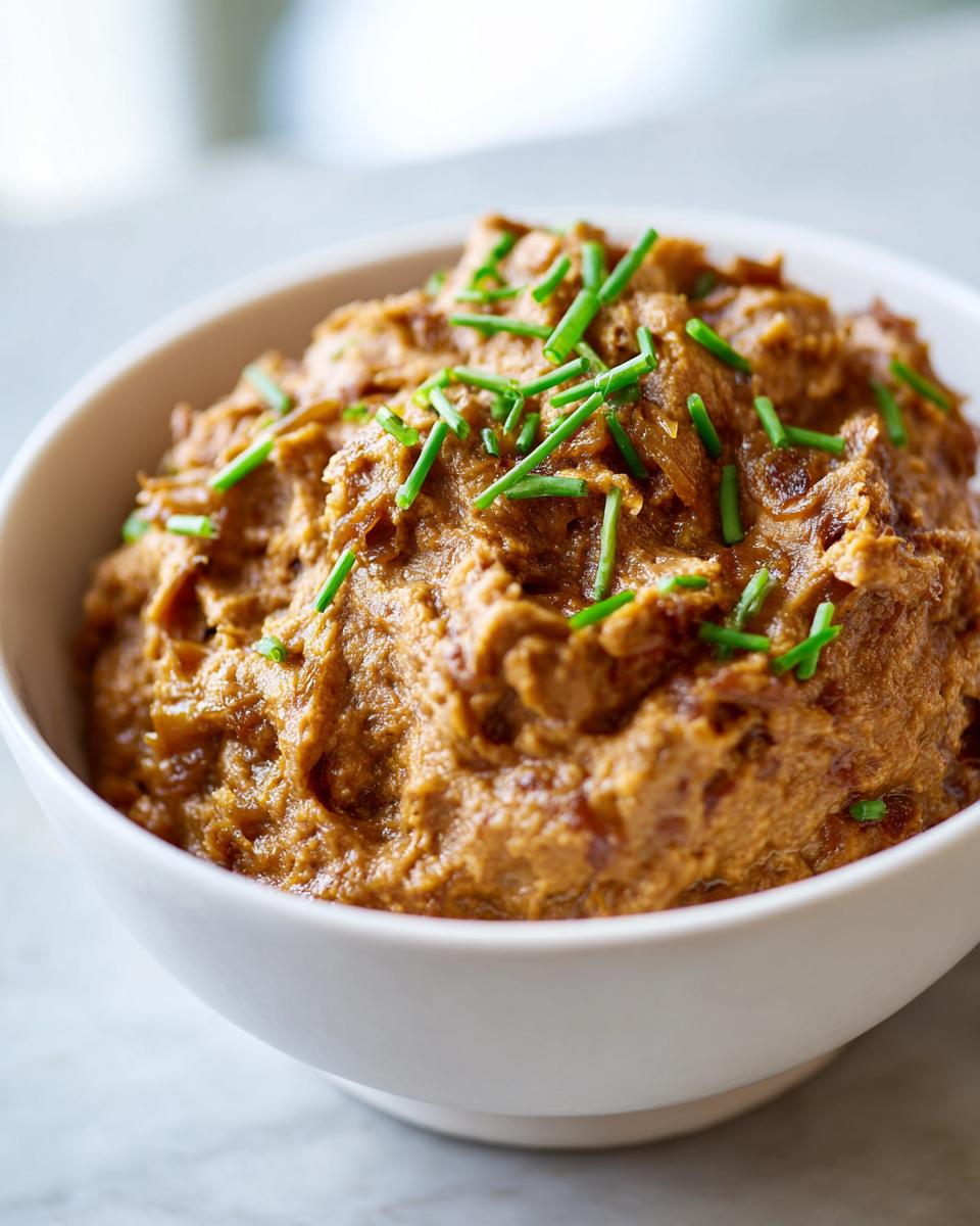 A close-up of thick, rich caramelized onion dip in a white bowl, garnished with fresh green chives.