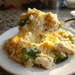 A spoonful of creamy, cheesy rice casserole with broccoli and chicken being lifted from a plate.