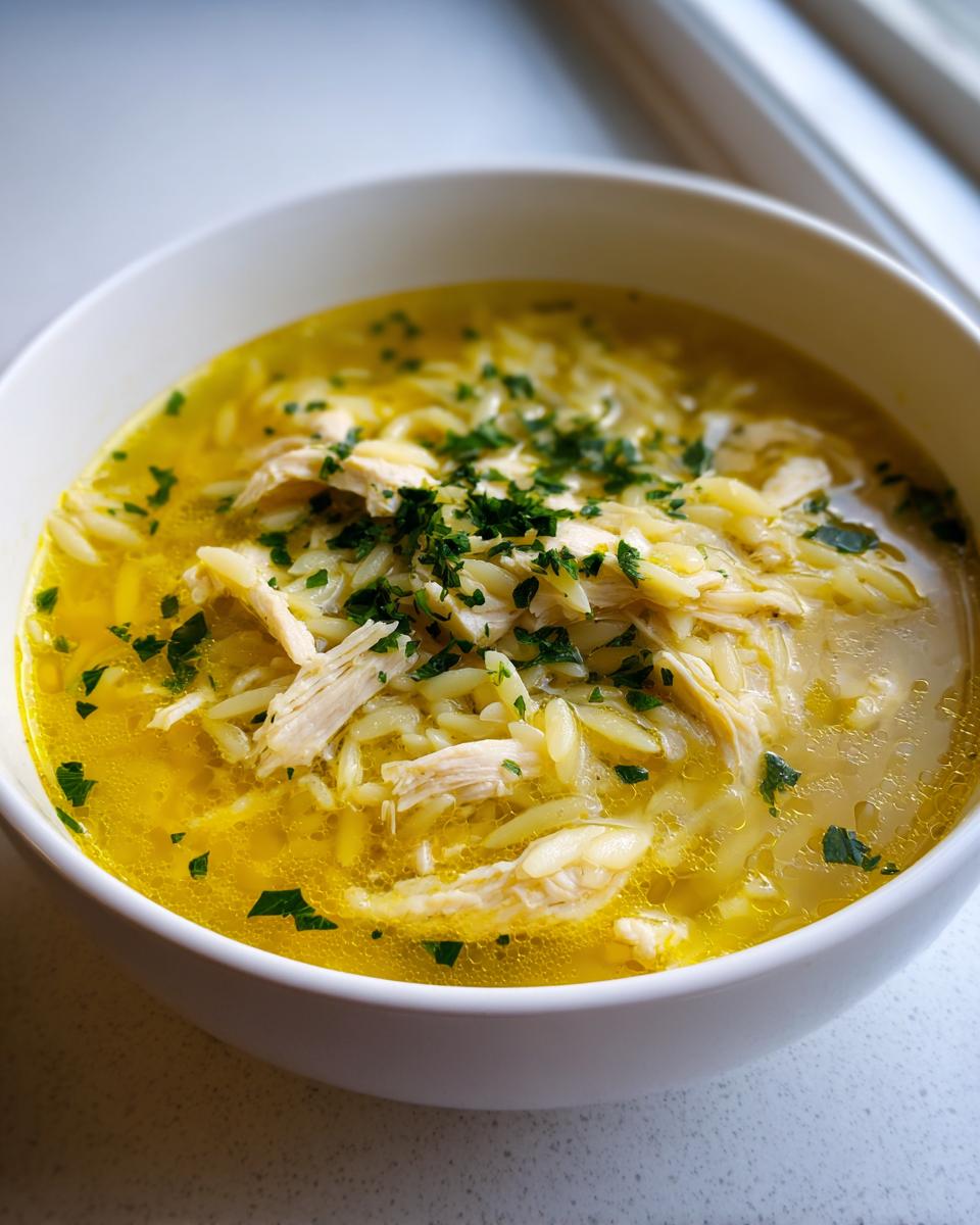 Close-up of a white bowl filled with bright yellow chicken orzo soup, featuring shredded chicken and topped with fresh parsley.