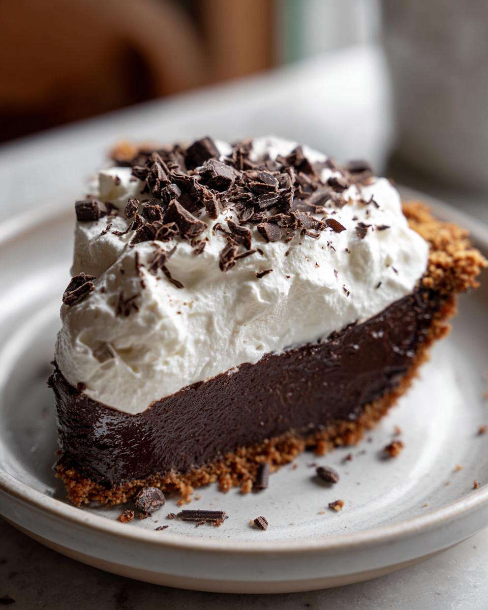 A decadent slice of chocolate cream pie with a thick, dark filling, graham cracker crust, and topped with whipped cream and chocolate shavings.