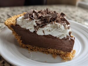 A decadent slice of chocolate cream pie with a graham cracker crust, topped with whipped cream and dark chocolate shavings.