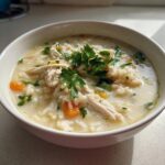 Close-up of a creamy bowl of chicken and rice soup, featuring shredded chicken, white rice, carrots, and fresh parsley garnish.