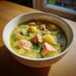 Close-up of a white bowl filled with creamy salmon soup, featuring chunks of pink salmon and yellow potatoes, garnished heavily with fresh dill.