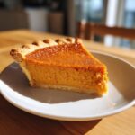 A perfect slice of creamy sweet potato pie with a flaky crust, sitting on a white plate in bright sunlight.